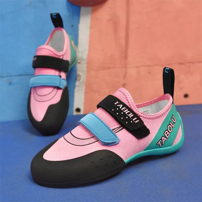 A pair of black and red beginner rock climbing shoes with a Velcro strap and wedge heel on a rocky background.