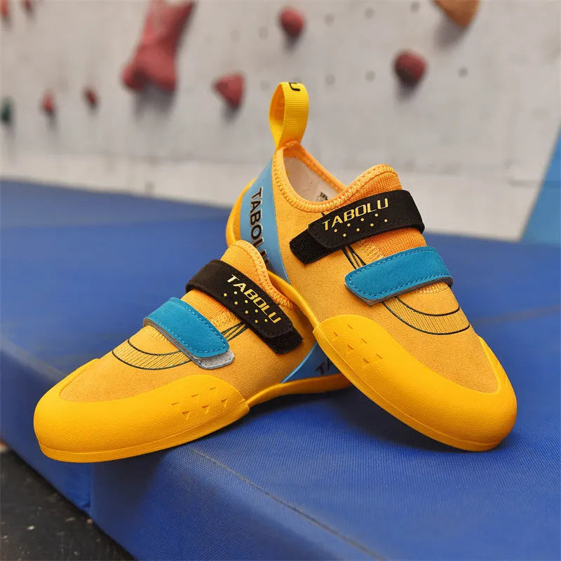 A pair of black and red beginner rock climbing shoes with a Velcro strap and wedge heel on a rocky background.