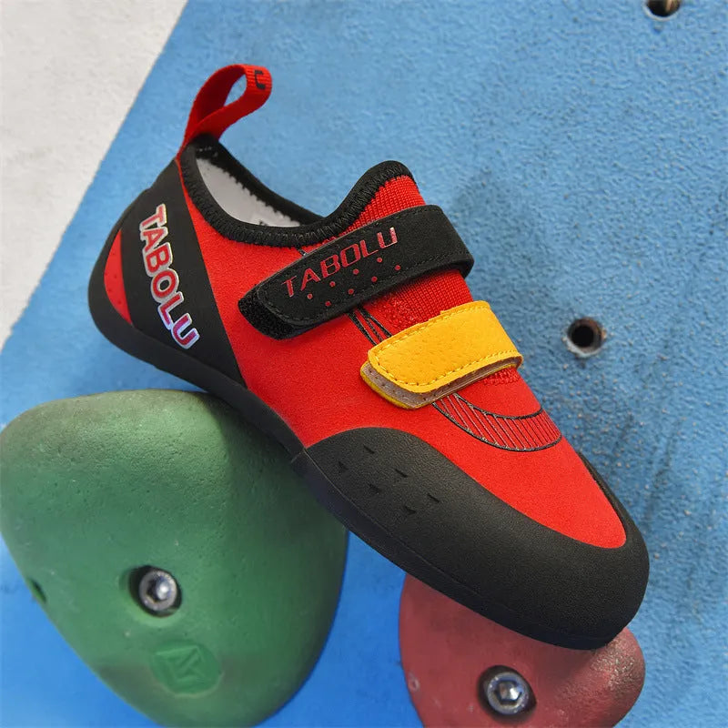 A pair of black and red beginner rock climbing shoes with a Velcro strap and wedge heel on a rocky background.