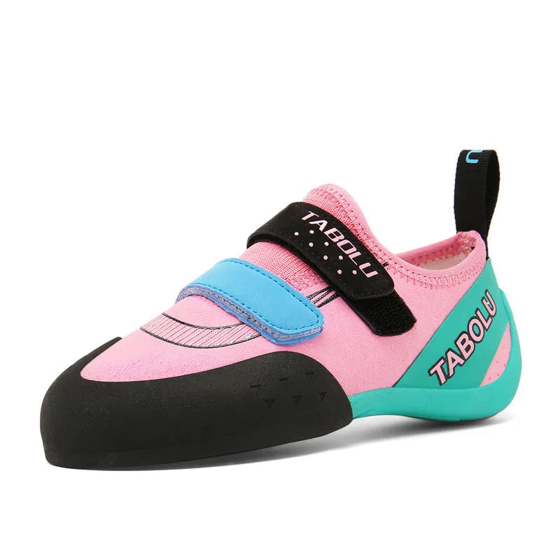 A pair of black and red beginner rock climbing shoes with a Velcro strap and wedge heel on a rocky background.